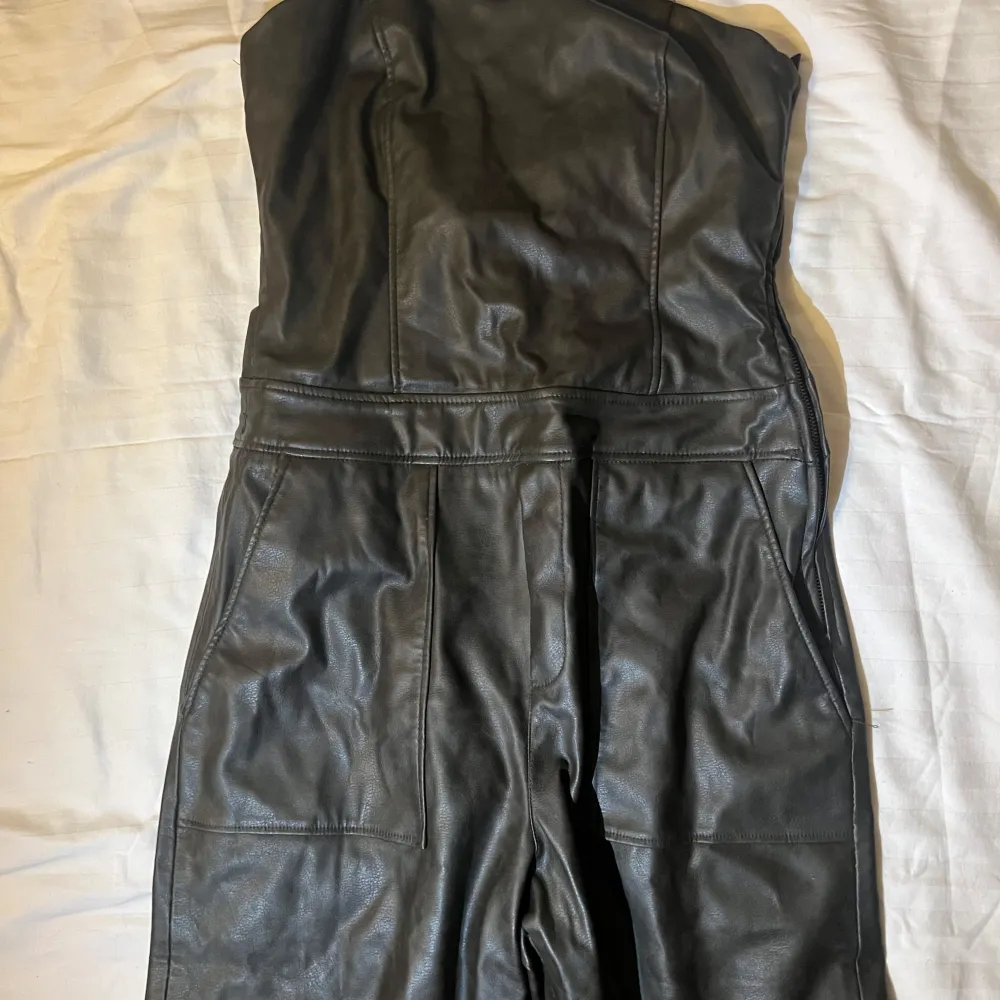Snygg svart axelbandslös jumpsuit från Zara i skinnimitation. Perfekt för en cool och stilren look. . Mekot.