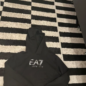 Svart hoodie från EA7 Emporio Armani - EA7 Tröja i Nyskick – Storlek 160 cm (12-15 år)  Säljer en stilren EA7-tröja i nyskick! Tröjan är perfekt för ungdomar och har storleken 160 cm, vilket passar för 12-15-åringar. Den är mycket välvårdad, utan några synliga tecken på användning