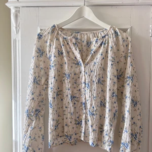 Blommig blus med långa ärmar - Säljer en superfin blommig blus i vitt med blå blommor. Den har långa ärmar och knappar framtill. Perfekt för vår och sommar, och den ger en romantisk och fräsch look. Passar både till vardags och festliga tillfällen!