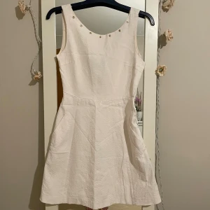 Vit klänning från Zara Basic - Säljer en superfin vit klänning från Zara Basic i storlek XS. Klänningen är ärmlös och har en stilren design med små nitar vid halsringningen. Perfekt för sommarens alla fester! Materialet känns lätt och bekvämt, och den har en dragkedja på sidan för enkel påklädning.