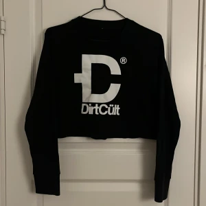 Svart croppad tröja från DirtCult - Säljer en svart croppad tröja från DirtCult med stort vitt tryck på framsidan. Tröjan har långa ärmar och är perfekt för en avslappnad stil. Fint skick då den knappt är använd. Perfekt för höst och vår!