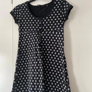 Svart kortärmad klänning med dödskallemönster från F.L.Y. - Säljer en cool svart klänning från F.L.Y. med ett unikt vitt dödskallemönster. Klänningen har korta ärmar och knappar framtill för en extra detalj. Perfekt för en avslappnad dag eller en fest med lite edge! 🖤