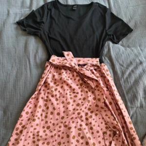 Svart och rosa klänning från Shein - Säljer en superfin klänning från Shein i storlek S. Den har en svart, ribbad överdel med korta ärmar och en rosa kjol med bruna prickar. Kjolen har ett knytband i midjan för en snygg passform. Perfekt för sommarens fester eller en dag på stan! 