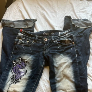 Blå jeans med fjärilsbroderi - Säljer ett par coola blå jeans med unikt fjärilsbroderi i lila och svart på både fram- och baksidan. Jeansen har en sliten look med ljusare partier och snygga detaljer som knappar och dragkedja. Perfekta för en avslappnad stil med en twist! passar inte mig som har M i jeans skulle säga att de är XS/S