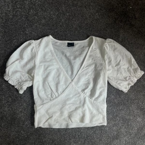 Vit omlottopp med puffärmar - Säljer en superfin vit omlottopp med puffiga kortärmar. Den är gjord i ett mjukt bomullsmaterial och har en v-ringad design som ger en snygg look. Perfekt för sommardagar eller en avslappnad festkväll!