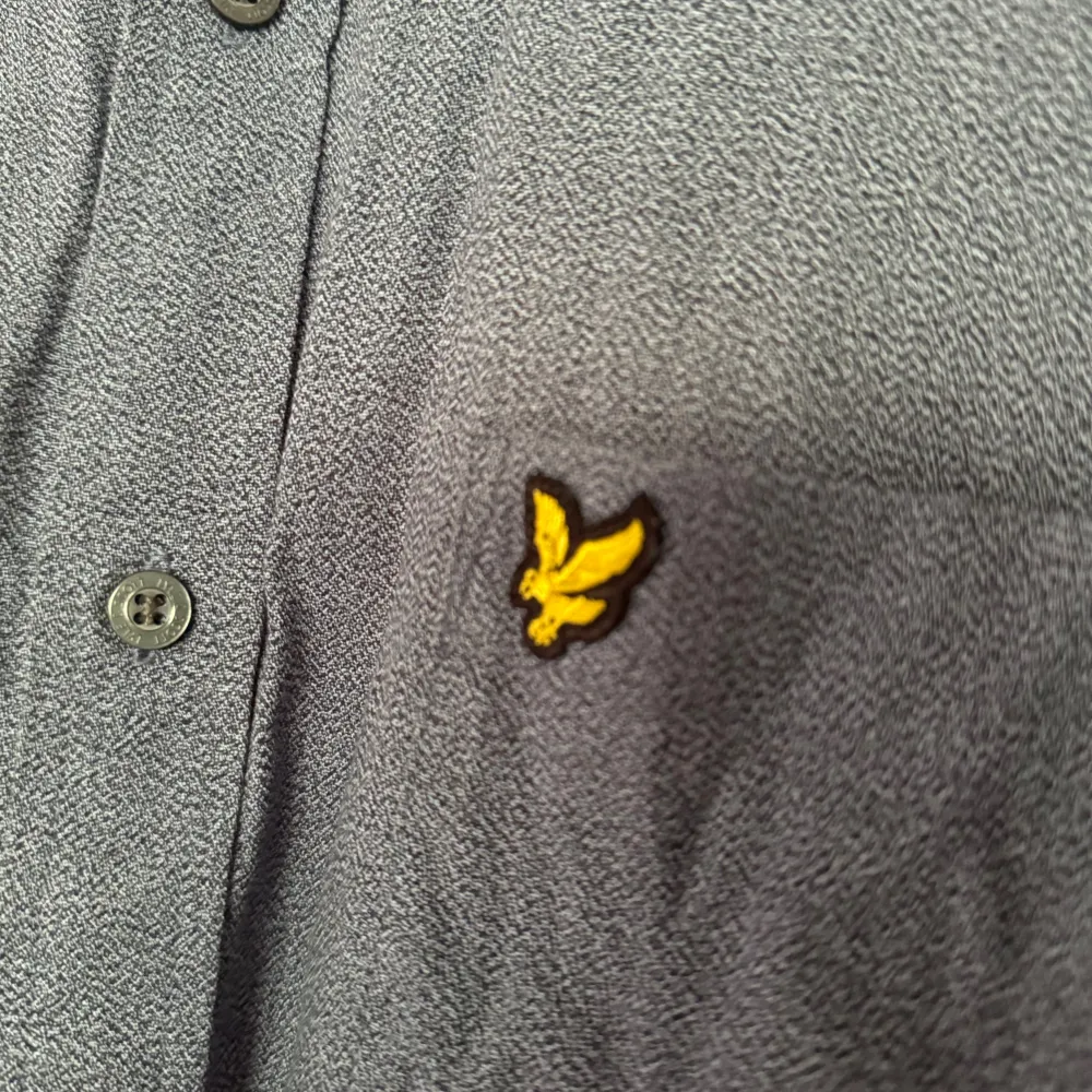 Säljer en snygg blå skjorta från Lyle & Scott. Den har en klassisk krage och långa ärmar, perfekt för både vardag och fest. Skjortan är dekorerad med det ikoniska gula logotypen på bröstet och har knappar hela vägen ner. Materialet känns bekvämt och stilrent.. Skjortor.