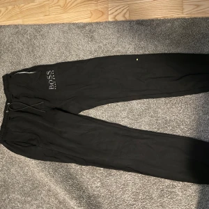 Svarta mjukisbyxor från Boss - Säljer ett par svarta mjukisbyxor från Boss. De har en bekväm passform med snörning i midjan och är perfekta för en avslappnad stil. Byxorna är i bra skick och har en stilren design med Boss-loggan på sidan.