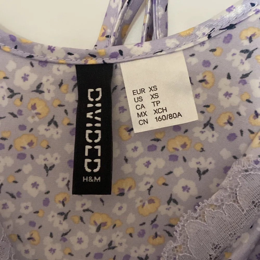 Säljer en superfin ljuslila klänning från H&M Divided i storlek XS. Den har ett sött blommigt mönster i gult och vitt, med spetsdetaljer vid halsringningen. Klänningen är ärmlös och har smala axelband, perfekt för sommarens fester eller en mysig vårdag. Materialet känns mjukt och skönt mot huden.. Klänningar.