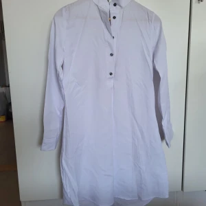 Skjorta  - Vit skjorta med långa ärmar och knappar framtill. Den har en liten krage. 