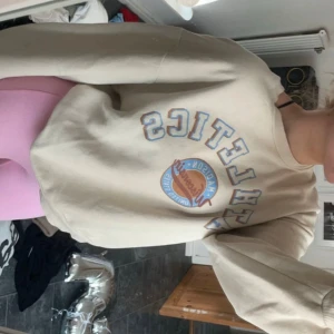 Beige sweatshirt med tryck - Säljer en supermysig beige sweatshirt med texten 'ATHLETICS' och ett coolt tryck i mitten. Perfekt för en avslappnad stil och passar till allt! Den har långa ärmar och en normal passform. Den är jättemysig och skön. Det kan förekomma defekter på den men inga märkvärdiga då jag inte ser några med blotta ögat.