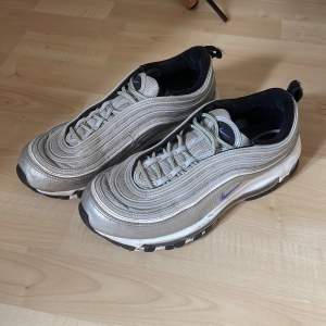 Säljer ett par Nike Air Max 97 i en cool silverfärg. Skorna har en futuristisk design med vågiga linjer och en synlig Air-enhet i sulan för extra komfort. De har en snygg lila detalj på sidan och svart insida. Perfekta för både vardag och fest!