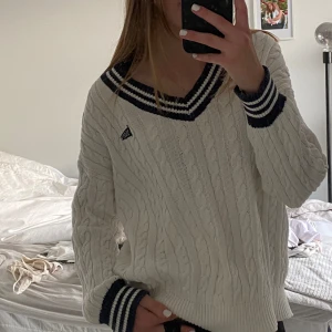 Vit kabelstickad tröja med marinblå detaljer - Säljer en superfin vit kabelstickad tröja med marinblå ränder vid krage och ärmslut. Tröjan har en v-ringad design och är perfekt för både höst och vinter. Den är långärmad och riktigt mysig att ha på sig. Perfekt för en casual look eller när du vill känna dig extra bekväm. (min kompis är modell)