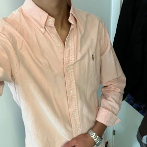 Ljusrosa skjorta från Ralph Lauren - Säljer en snygg ljusrosa skjorta från Ralph Lauren i storlek S. Skjortan är långärmad och har klassiska knappar framtill. Den är gjord av bomull och har den ikoniska broderade loggan på bröstet. Perfekt för både vardag och fest! Den är äkta!