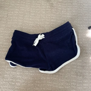 Marinblåa shorts från Divided - Säljer ett par svarta shorts från Divided H&M i storlek S. De är gjorda av en mjuk blandning av bomull och polyester, vilket gör dem superbekväma. Shortsen har en vit kant och en knytning i midjan för en justerbar passform. Perfekta för en avslappnad dag eller träning!