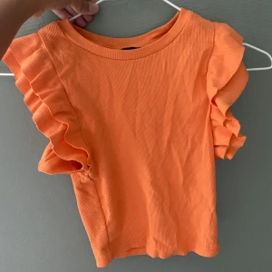 Orange ribbad topp med volangärmar - Säljer en superfin orange ribbad topp med volangdetaljer på ärmarna. Perfekt för sommardagar eller en festlig kväll. Toppen är i ett mjukt och stretchigt material som sitter bekvämt.