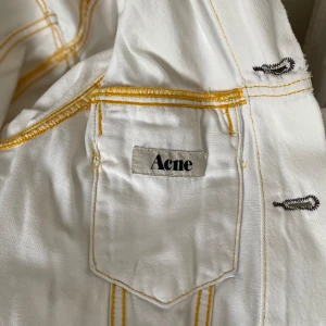Vit jeansjacka från Acne - Säljer en snygg vit jeansjacka från Acne. Den har en klassisk design med brons knappar och kontrasterande sömmar i gult. Jackan har två bröstfickor med lock och knappdetaljer. Perfekt för vår och sommar! Säljer pga knappt använd.
