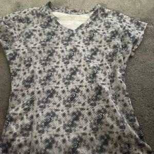 Mönstrad t-shirt från 2XU - Säljer en cool t-shirt med ett unikt svartvitt mönster. Den har en v-ringad hals och korta ärmar, perfekt för träning eller en avslappnad dag. Materialet känns lätt och andas bra, vilket gör den idealisk för aktiva dagar.