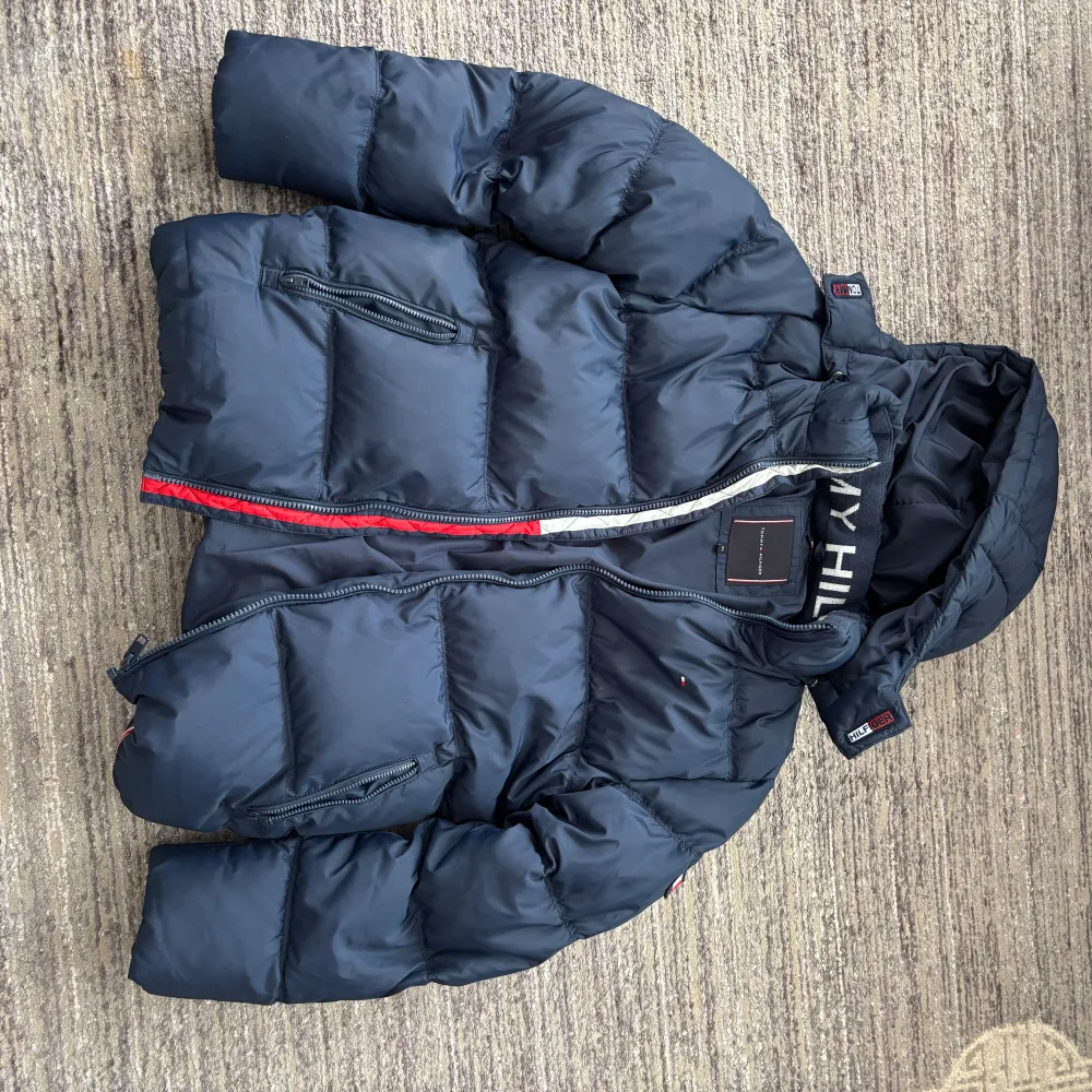 Säljer en snygg och varm mörkblå dunjacka från Tommy Hilfiger. Jackan har en praktisk huva och dragkedja framtill. Perfekt för kalla vinterdagar. Den är i bra skick och har en stilren design med märkets logga på bröstet.. Jackor.