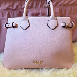 Rosa handväska från Burberry - Säljer en superfin rosa handväska från Burberry. Den har ett klassiskt rutigt mönster på sidorna och är gjord i skinn. Väskan har två handtag och snygga spänndetaljer på sidorna. Insidan är rymlig med flera fack och en dragkedja för säker förvaring. Perfekt för både vardag och fest!