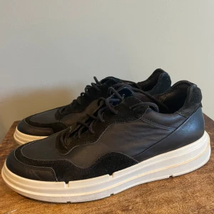 Svarta sneakers från Ecco - Snygga svarta sneakers från Ecco, skinn med detaljer i mocka med vit sula som ger en cool kontrast. Perfekta för både vardag och lite mer uppklädda tillfällen. Skorna har snörning och är knappt använda även om sulan är lite smutsig. Dyra i inköp. 