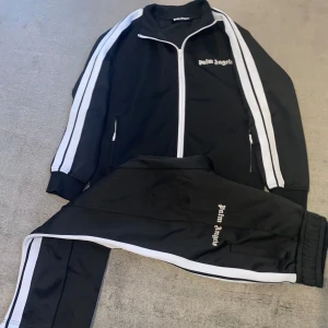 Svart träningsjacka från Palm Angels - Säljer en svart träningsjacka från Palm Angels med vita ränder längs ärmarna. Jackan har dragkedja och är långärmad, perfekt för både träning och chill, superbekväm!