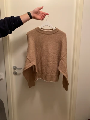 Beige stickad tröja - Halvhög krage. Vit kant i kragen, armslut och mudd. Nedhasade axlar. Loose fit
