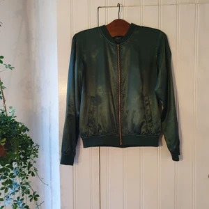 Jacka grön skimrig storlek M - Jacka i grönt och skimrigt/glansigt material. Dragkedja i guldaktig ton. Tunt material. Knappt använd och i god kvalité.