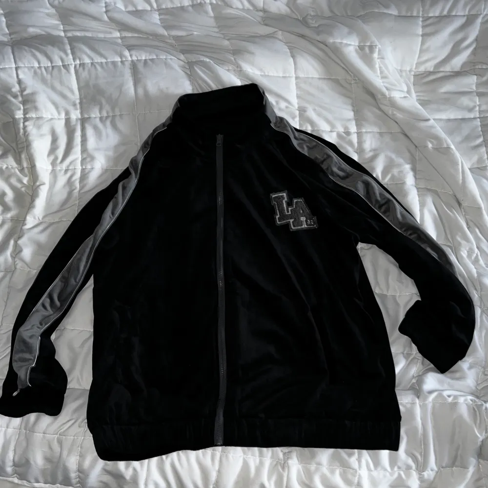 En svart tröja med mörkgråa detaljer och grått tryck i tyg som står LA. Är något mjukare material som är riktigt skönt. Storlek L . Hoodies.