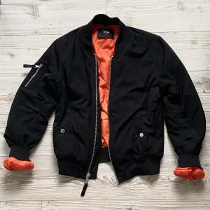 Bomberjacka <3 - Ball svart bomberjacka med innertyg i orange färg 🧡 Jackan har två fickor fram med knappar och en ficka på ena ärmen med dragkedja. Storlek XS men sitter lite oversized och passar XS/S/M. 