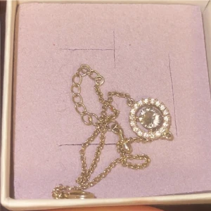 Lily and rose - Lily and rose armband. Det är svårt att se färgen på bild. Kedjan är guld, små stenarna är rosa och stenen i mitten är grå/grön. 