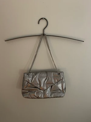 Silvrig My Mo Now Väska - Använd ca 5 gånger, i utseende oanvänd/nyköpt.  Materialet känns och ser ut som en dunjacka. Köpt för 1135kr.