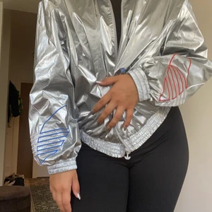Adidas windbreaker - Adidas jacka i supercool silvrig färg. Den är stor men sitter ändå åt i midjan när man har den stängd. Har adidas loggan på ärmarna i blått och rött