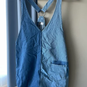 Jeans klänning  - Hängselklänning i jeanstyg. Tyg i två olika nyanser och fransig ”avklippt” nederkant . Kort, vid modell. 