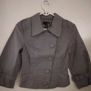 HELT NY & OANVÄND grå vårjacka / sommarjacka. Strlk: 34/XS. Felfri. - HELT NY & OANVÄND grå kort vårjacka / sommarjacka med medveten sned knäppning. Strlk: 34/XS. Material: Bomull.  Stängs med knappar och har 2 fickor. Felfri.  FRAKT TILLKOMMER. 