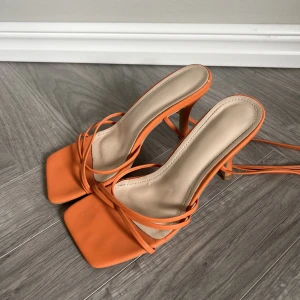 Orangea klackskor med långa band! - Orangea klackskor med långa band som går att knyta som man själv önskar! Superhärlig färg! Strl 36. Uppskattningsvis ca 7 cm hög klack. Har använts och har lite slitning vid hälen. Annars fint skick! 