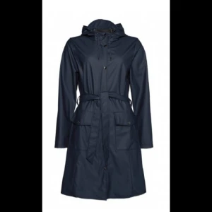 Regnjackan  - Denna regnjacka från Rains kommer i en snygg färg med en matt look. Regnjackan har knytband runt midjan, så jackan kan snöra in sig och ge en feminin passform. Regnjackan har dragkedja och snäpper ner hela vägen, två rymliga fickor fram