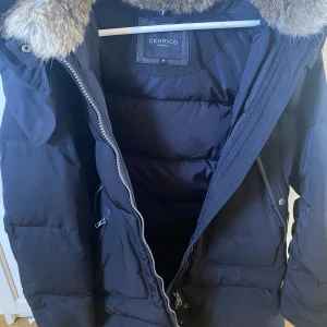 Cedrico Monet jacka - En Cedrico Monet jacka i svart. Storlek M. Varm och skön. Päls på kragen som är avtagbar, även päls i luvan, där hela luvan kan tas av.  Använd i fint skick, enda att anmärka på är att det saknas en knapp på spännerna längst ner. Syns på sista bilden.  