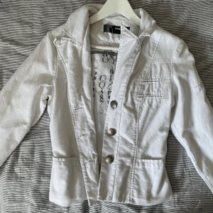 Tunnare jacka - Vit bomullsjacka som liknar en kavaj. Jättefina mönstrade knappar i metall. Påsydda och strukna ”patches” bak på ryggen! 🤍✨Spårbarfrakt på 59kr är inkluderat i priset!