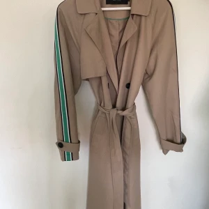 Trenchcoat - Trenchcoat med gröna ränder på ärmarna. Råkat bli ett streck av bläckpenna på framsidan, går säkert bort i tvätten. Stl L - oversized på mig som är XS/S! Pris 90kr