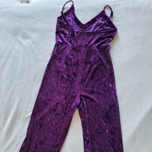 Lila jumpsuit i krossad sammet - Hellång jumpsuit i krossad sammet. Stretchingt material. Figurnnära topp och vidare ben. Fantastisk lila färg! Justerbara spaghetti-axelband och bälteshällor i sidorna. Från NA-KD.  95% polyester 5% elastan  Toppenskick!
