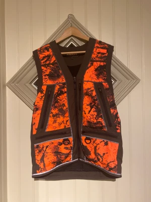 Härkila - Hundförarväst- Wildboar PRO Jaktväst som passar till hundförare till såväl passjägare med en rad funktionella funktioner. Fickor på framsida till både radio och för patroner även stor ficka på baksidan. Västen är oanvänd.