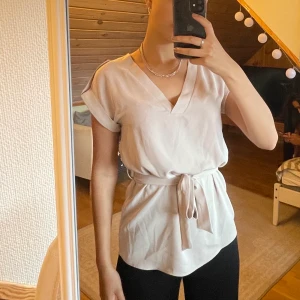 Ljusbeige blus med knytning - Blusen är använd fåtal gånger och i bra skick. Super fin med knappar i guld på axlarna och knytband i midjan.