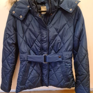 Jeane Blush jacka i nyskick - Blå Jeane Blush jacka med hög krage och huva i fuskpäls. Aldrig använd. Avtagbar huva samt skärp i midjan som går att ta av. Finns en innerficka på vänster sida samt två fickor framtill. Storlek S. Material i 100% polyester