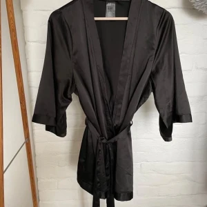 Svart kimono i sidenliknande material - Vad: Svart kimono i sidenliknande material. Skärp och fin detalj på ryggen.  Storlek: XS (passar även S) Märke: 97% polyester 3% elastan  Skick: Utan anmärkning 