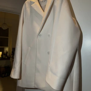 Kort vit kappa  - En kort vit kappa från NA-KD.   Skriv för fler bilder. Ett år gammal och använd fåtal gånger. (Ser lite missfärgad ut vid kragen pga kamerans kvalité). Endast en liten fläck som går bort i tvätten på kragen.   Pris kan diskuteras. Köparen står för frakt.🌸