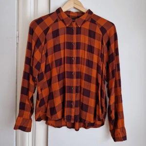 Rutig skjorta Monki - Skjorta i viscose.  Rutig i orange och vinröd. Små "slitsar" på sidorna.  Passar stl M / 40 / 42