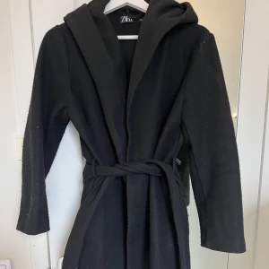 Svart kappa  - Säljer denna kappa från zara. Kappan har inga direkta defekter, förutom att materialet är lite nopprigt. Det är en luva på kappan och ett tillhörande skärp i midjan. 