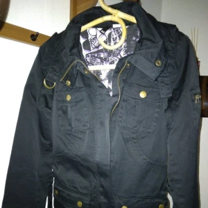 HELT NY & OANVÄND Snygg svart parkas jacka / damjacka. Storlek: 32/XS. - Helt NY och oanvänd snygg svart jacka i parkas-stil, för dam/tjej. Storlek: 32/XS. Material: Bomull. Stängs med dragkedja, avtagbart skärp i midjan , avtagbar luva, snyggt svart-vit mönstrat foder inuti, samt flera fickor. Helt felfri. FRAKT TILLKOMMER.