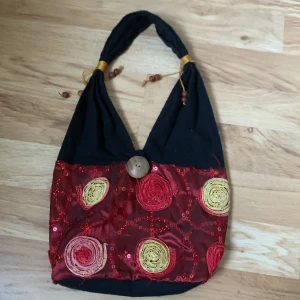 Handväska  - Gullig liten handväska med handbroderade blommor och paljetter. Stängs med en träknapp. Handtaget är prytt med gulligt snöre och trapärlor. Väskan har en strukturerad botten och är 100% bomull.