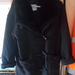 Ullkappa kort stl L. Svart/grå. - Fin ullkappa kort med syntetiskt pälskrage knäpoes med knappar .finnes ett litet hålfram. Fickor fram ,fodrad. Storlek L. Hämtas i Skene alt skickas .